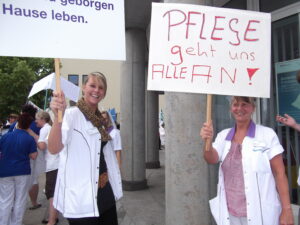 Zu sehen sind zwei Frauen in einem weißen Kittel. Sie halten Schilde hoch und lächeln in die Kamera. Auf dem einen Schild steht Pflege geht uns alle an!